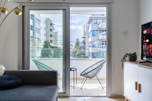 A modern living room with a sofa, a chair, a TV, and a sliding glass door leading to a balcony with a view of other buildings. - Classy 2BR Cafe Suite Strovolos (Nicosia)