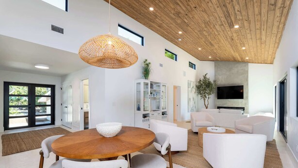 Uma sala de estar moderna com uma mesa de jantar redonda de madeira, sofás brancos e uma luminária grande de fibras trançadas.