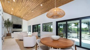 Uma sala de estar moderna com teto de madeira, sofás brancos, uma mesa redonda de madeira e grandes janelas com vista para o exterior.