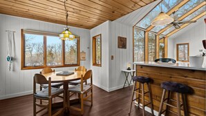 A cozy cabin interior with a wooden dining table, chairs, a bar area, and large windows offering views of the outdoors.
