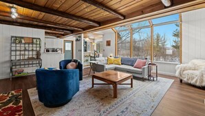 A cozy living room with a wooden ceiling, a blue armchair, a grey sofa, and a large window offering a view of trees.