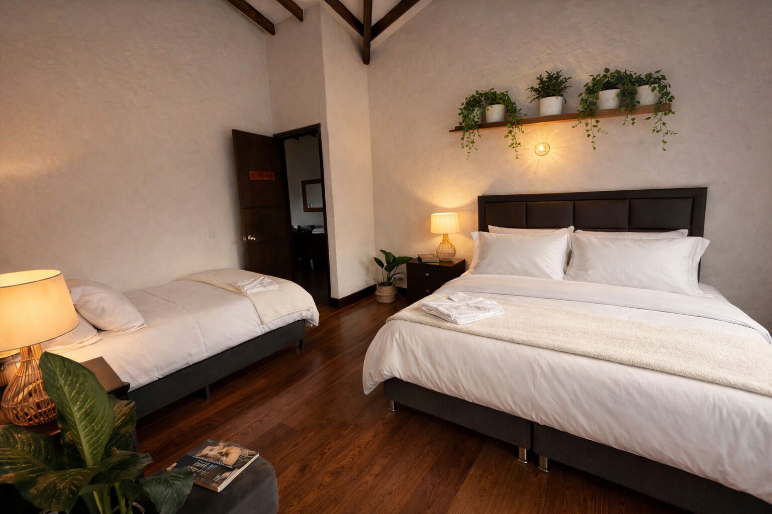 A hotel room with two beds, a nightstand with a lamp, a shelf with plants, and a wooden floor.