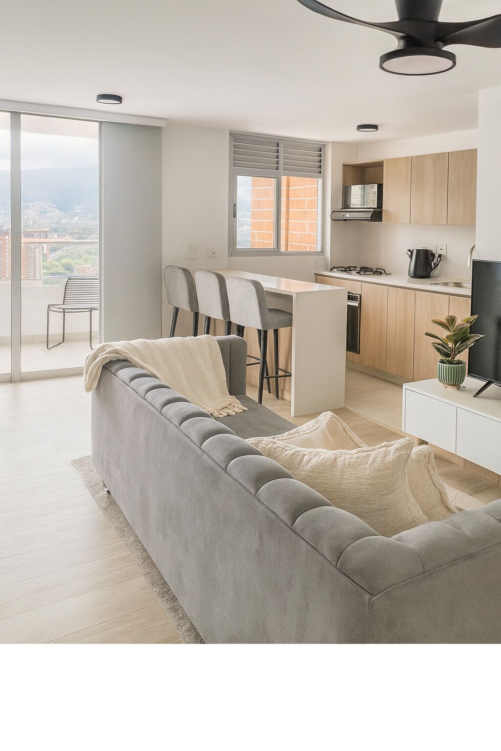 Un apartamento moderno con sofá gris, una pequeña cocina y un balcón con vista a la ciudad.