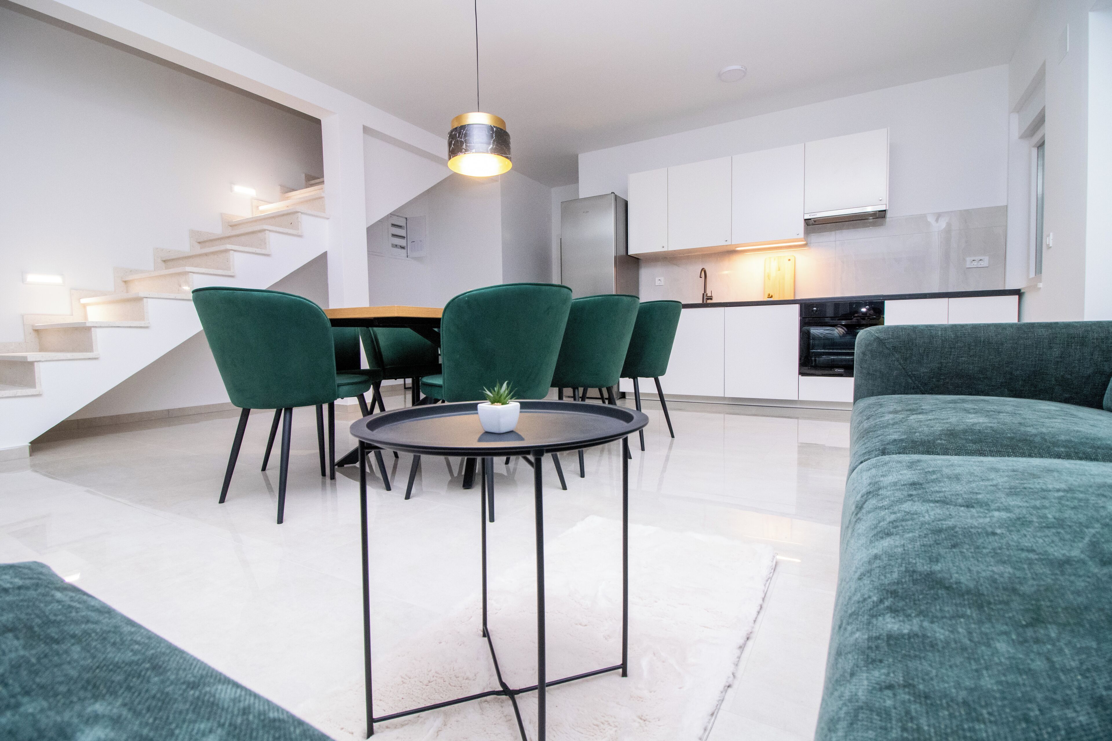 A modern living room with a round table and green chairs, a staircase, and a kitchen area with white cabinets and a stainless steel refrigerator.