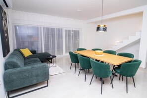 Een moderne woonkamer met een modulaire bank, een eettafel met stoelen, een trap en een groot raam met dunne gordijnen.