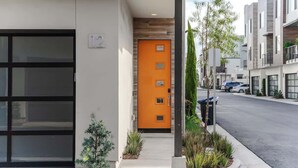 Un bâtiment moderne avec une porte orange vif, une grande fenêtre et un petit arbre au premier plan.