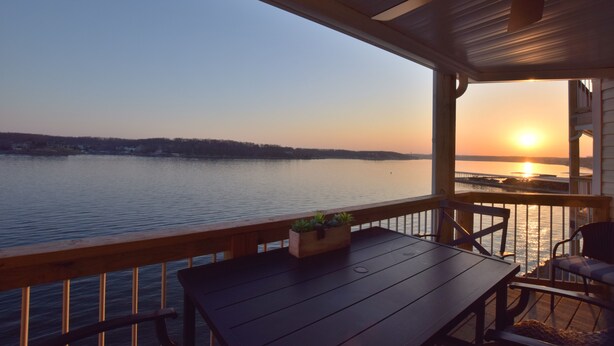 Una terraza de madera con una mesa y sillas, con vista a un lago tranquilo al atardecer.