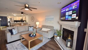 Una sala de estar con chimenea, un televisor mostrando servicios de transmisión y una cocina al fondo.