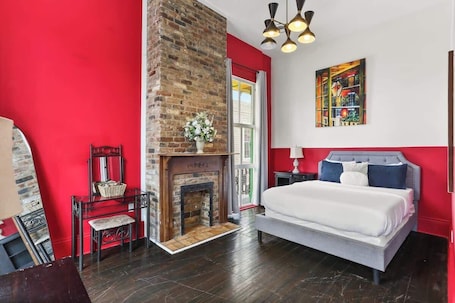A bedroom with a brick fireplace, a bed with a grey headboard, a nightstand, and a painting on the wall.