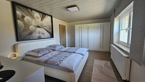 Een slaapkamer met een bed, nachtkastje en een groot abstract schilderij. Er is een raam met zonwering en een radiator.
