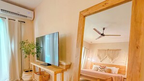 Een moderne woonkamer met een houten vloer, een grote spiegel, een televisie op een houten dressoir, een plafondventilator en een bed met hoofdeinde op de achtergrond.