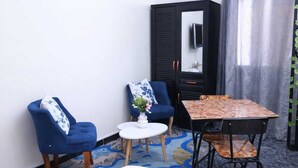 Una habitación pequeña con una cama, dos sillones azules, una mesita redonda con un florero y una mesa de madera con sillas.
