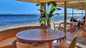 A wooden deck with a round table and chairs, overlooking a beach with a clear view of the sea and distant mountains. - Exotic escape - luminous, charming veranda (Bormes-les-Mimosas)