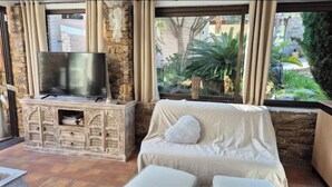 A living room with a stone fireplace, a flat-screen TV, a beige sofa, and a view of a garden through large windows. - Exotic escape - luminous, charming veranda (Bormes-les-Mimosas)