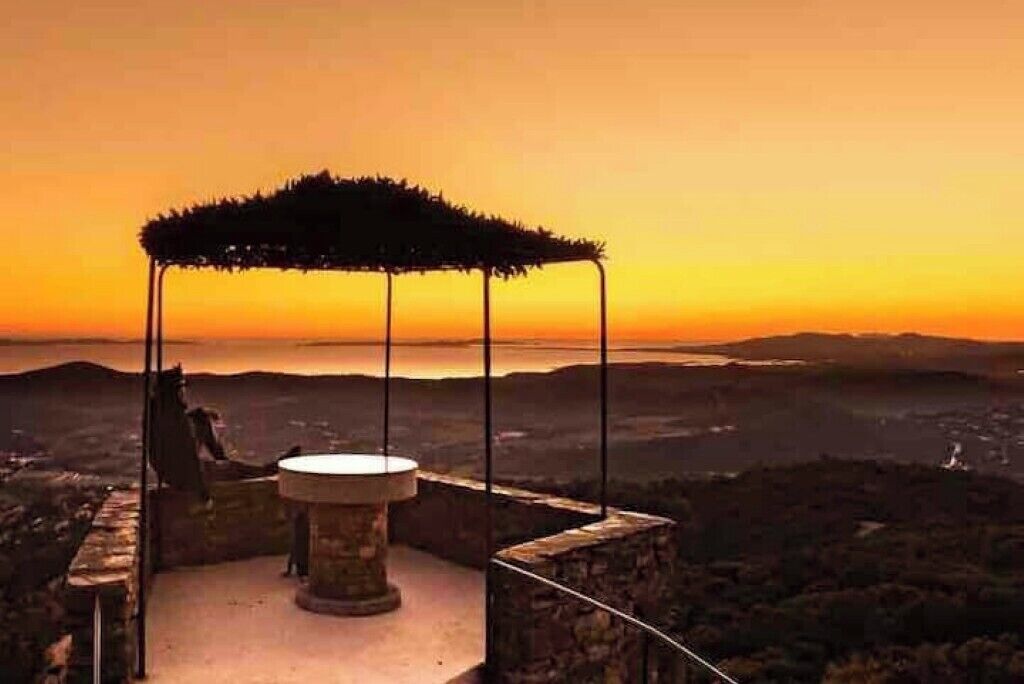 Ein Steingebäude mit einem runden Tisch und einem Reetdach, mit Blick auf eine Landschaft mit Hügeln und einem Gewässer bei Sonnenuntergang.