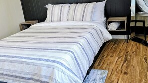 Un dormitorio con una cama grande, suelo de madera, cabecera oscura y una mesita de noche con lámpara.
