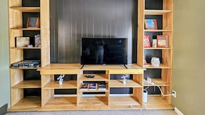 Un mueble de madera con un televisor de pantalla plana, repisas con juegos de mesa y un enchufe de corriente blanco.