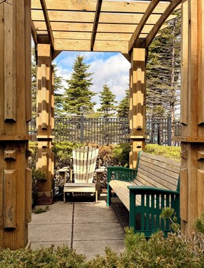 Puinen pergola, jonka alla on penkki ja tuoleja. Pergolaa ympäröi vihreä kasvillisuus ja aita.