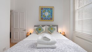 A neatly made bed with a floral-patterned bedspread, two matching pillows, and a folded white towel. A colorful artwork hangs above the bed, and a window with blinds is visible to the right. - Quirky Hampstead Flat RH6 (London)