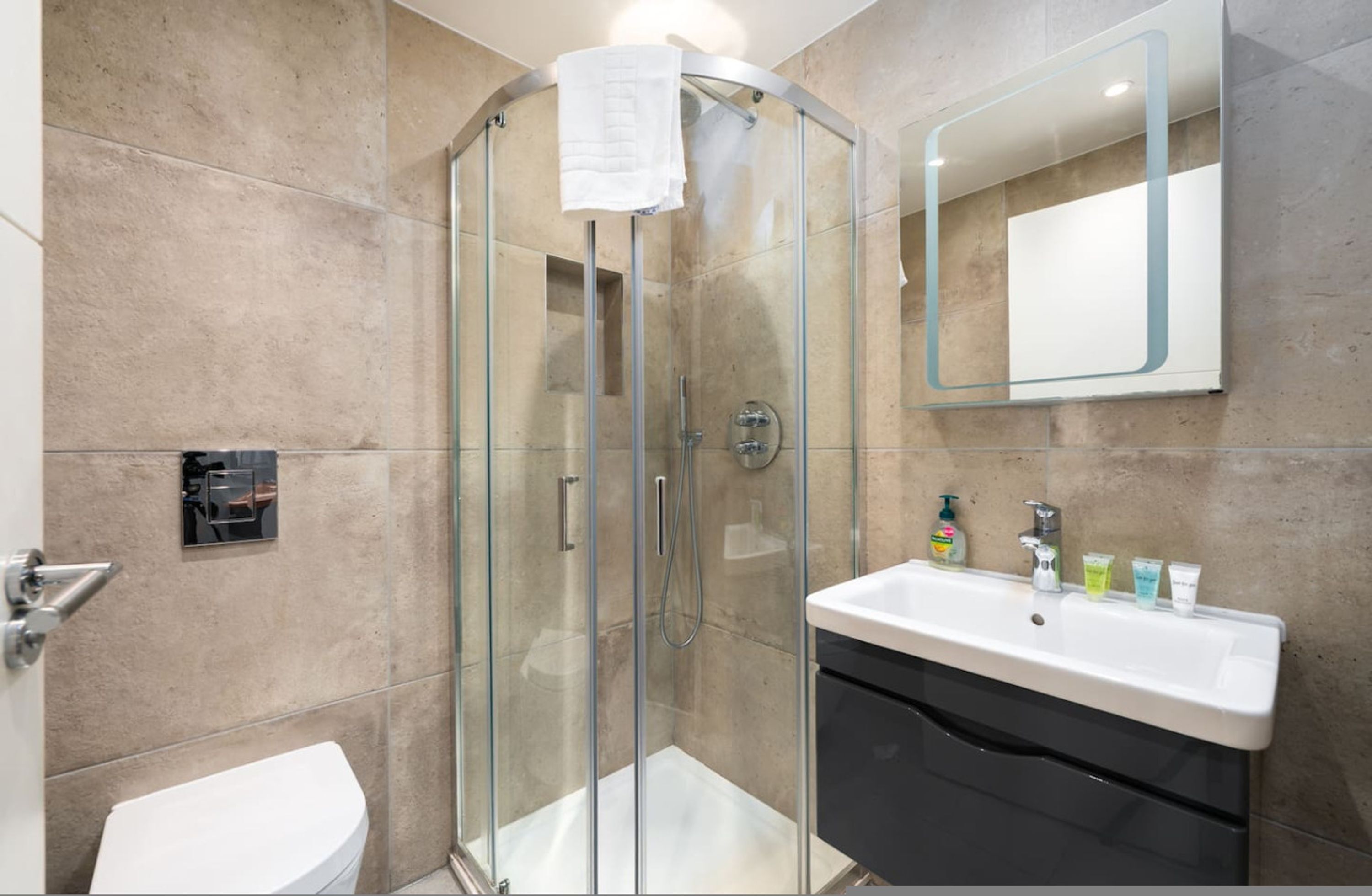 Un baño moderno con ducha de vidrio, lavamanos blanco con artículos de aseo y un espejo grande.