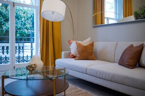 Ein Wohnzimmer mit einer Couch, einem Glastisch, einer Stehlampe und Blick durch ein Fenster mit Vorhängen.