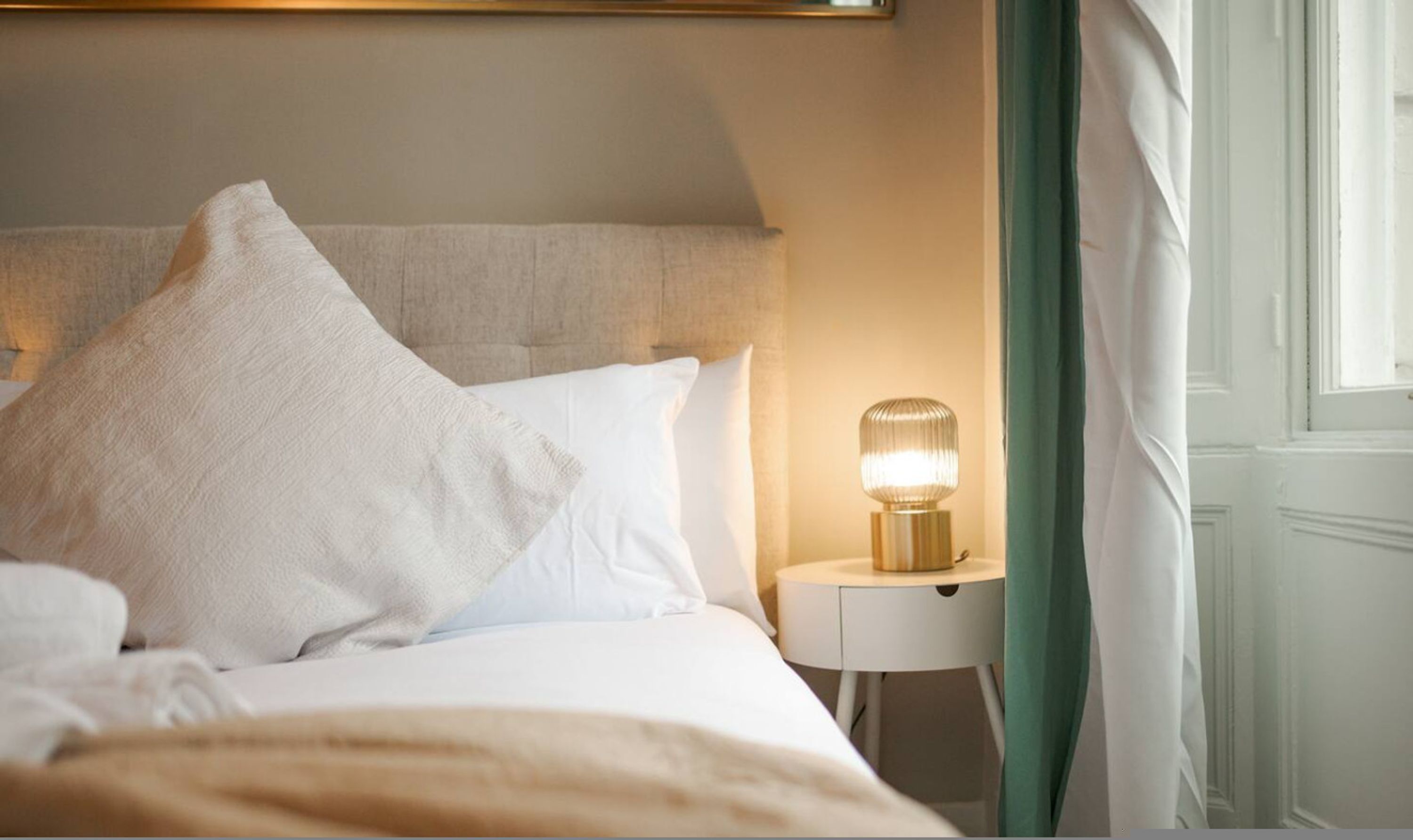 A neatly made bed with white linens and pillows, a bedside table with a lamp, and a view of a window with curtains.