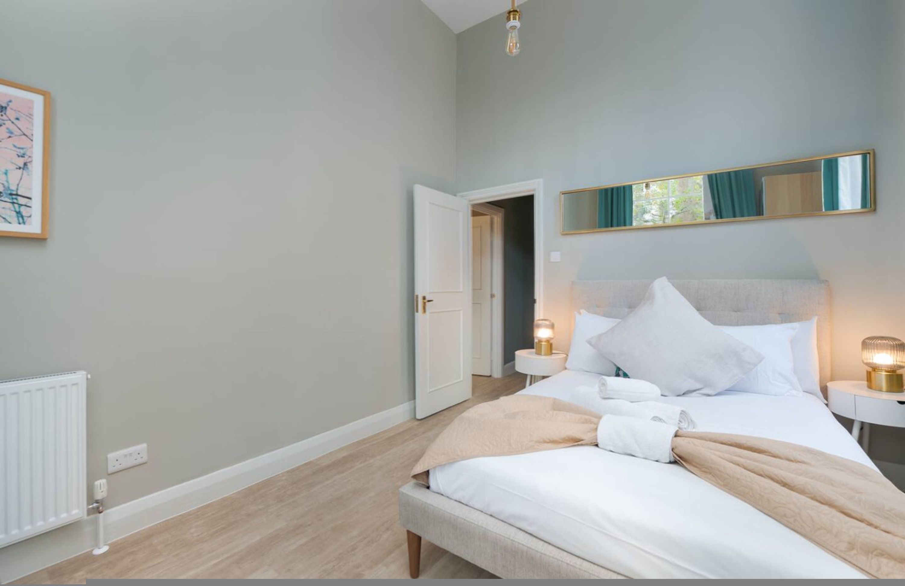 A bedroom with a bed, bedside lamps, a mirror, and a door leading to another room.
