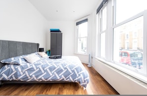 A modern bedroom with a large bed, grey wardrobe, and a view of the street through large windows. - Gorgeous Apartment by the Park SS2 (London)
