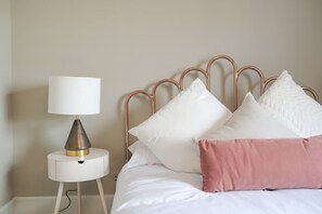 Un lit bien fait, avec une literie blanche et un oreiller rose, une table de chevet blanche avec une lampe, et une tête de lit en métal.