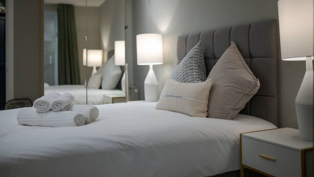 A neatly made bed with rolled towels, a grey upholstered headboard, and bedside lamps. - Stylish Hyde Park Apartment PS2 (London)