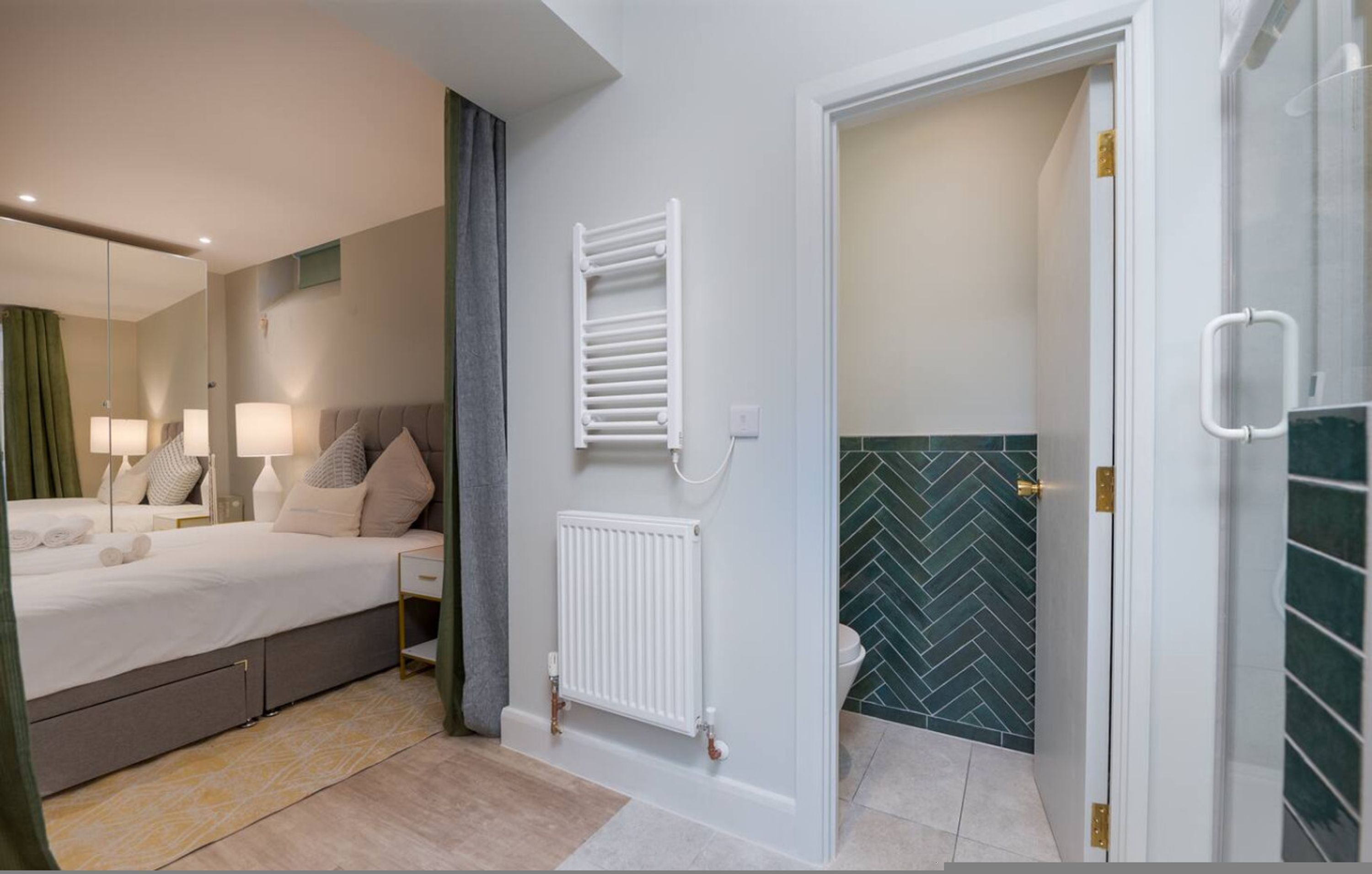 A modern hotel room with a bed, bedside lamps, a radiator, and a shower area with a herringbone tile backsplash.