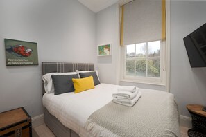Een net opgemaakt bed met een deken in wit en grijs, een geel kussen en een opgevouwen witte handdoek. Naast het bed staat een klein houten tafeltje, een grijs hoofdbord en een raam met een gele gordijn.