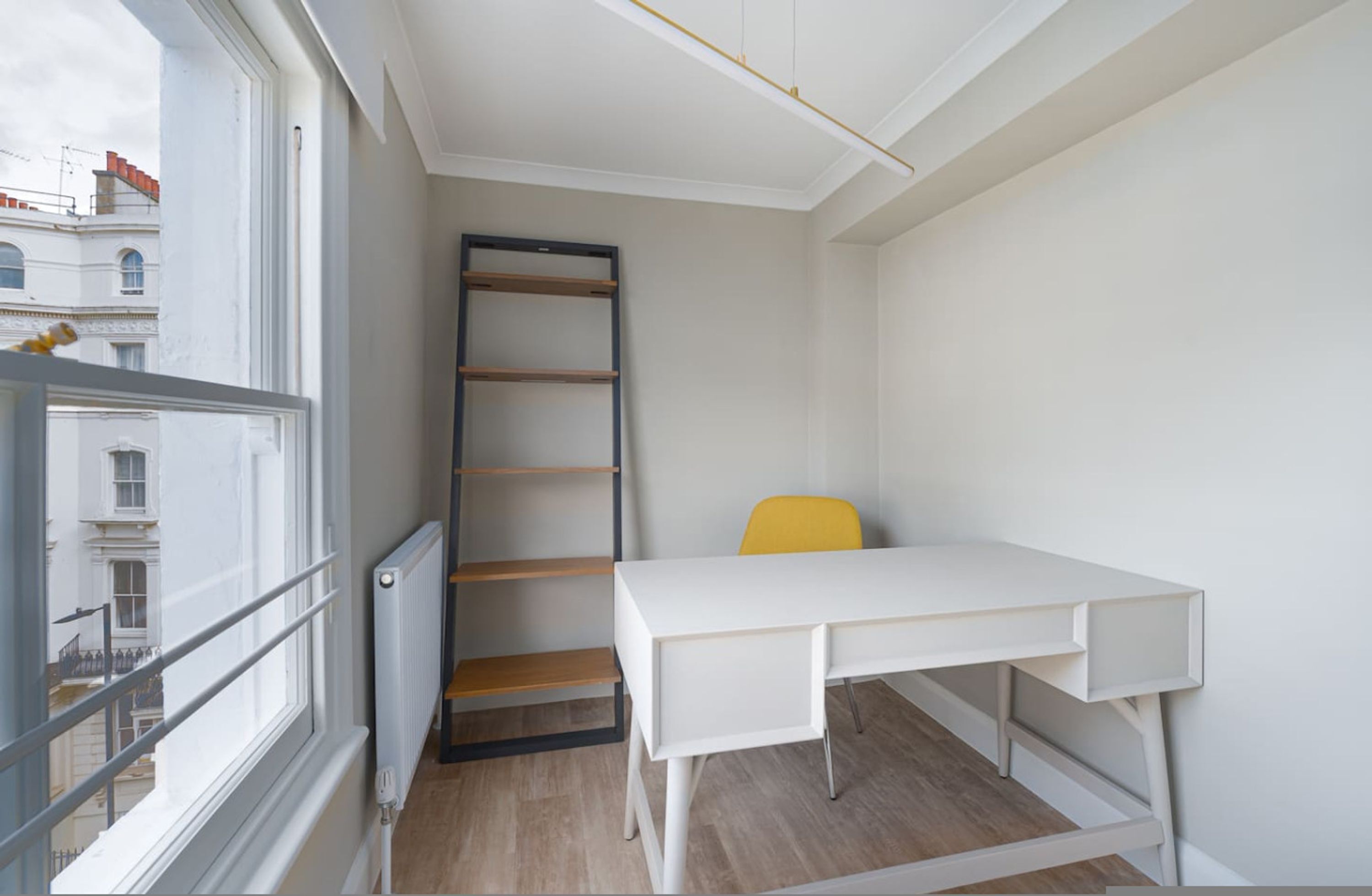 Une pièce avec un bureau blanc, une chaise jaune, une échelle en bois et une fenêtre donnant sur des immeubles.