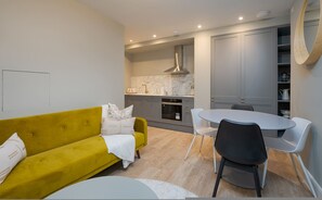 Un salon moderne avec un canapé jaune, une table à manger avec des chaises et un coin cuisine doté de meubles gris et d’un dosseret en marbre.