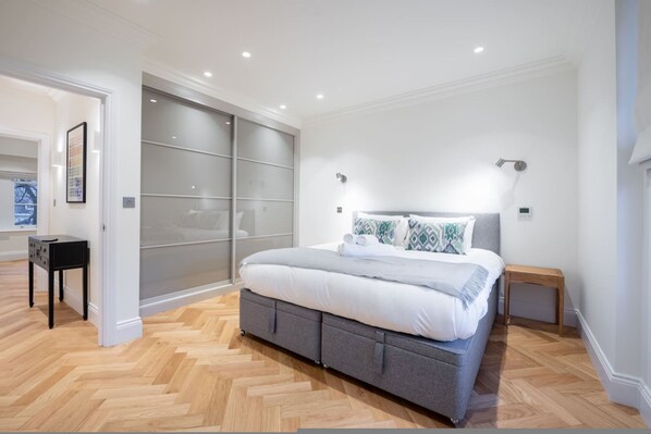 A modern bedroom with a large bed, a sliding glass door closet, and a small wooden nightstand. - Beautiful Hampstead Home RH9 (London)