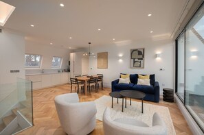 A modern living room with a blue sofa, white armchairs, and a dining table. There is a glass railing on the left and a large window on the right. - Beautiful Hampstead Home RH9 (London)
