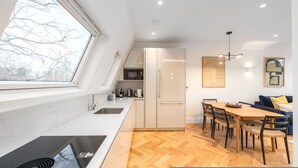 A modern kitchen with a large window, a dining area with a wooden table and chairs, and a built-in oven. - Beautiful Hampstead Home RH9 (London)