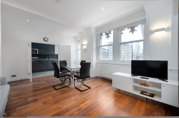 A modern living room with a wooden floor, a dining area with a glass table and chairs, a TV on a white stand, and a kitchen in the background. - Beautiful London Flat FS3 (London)