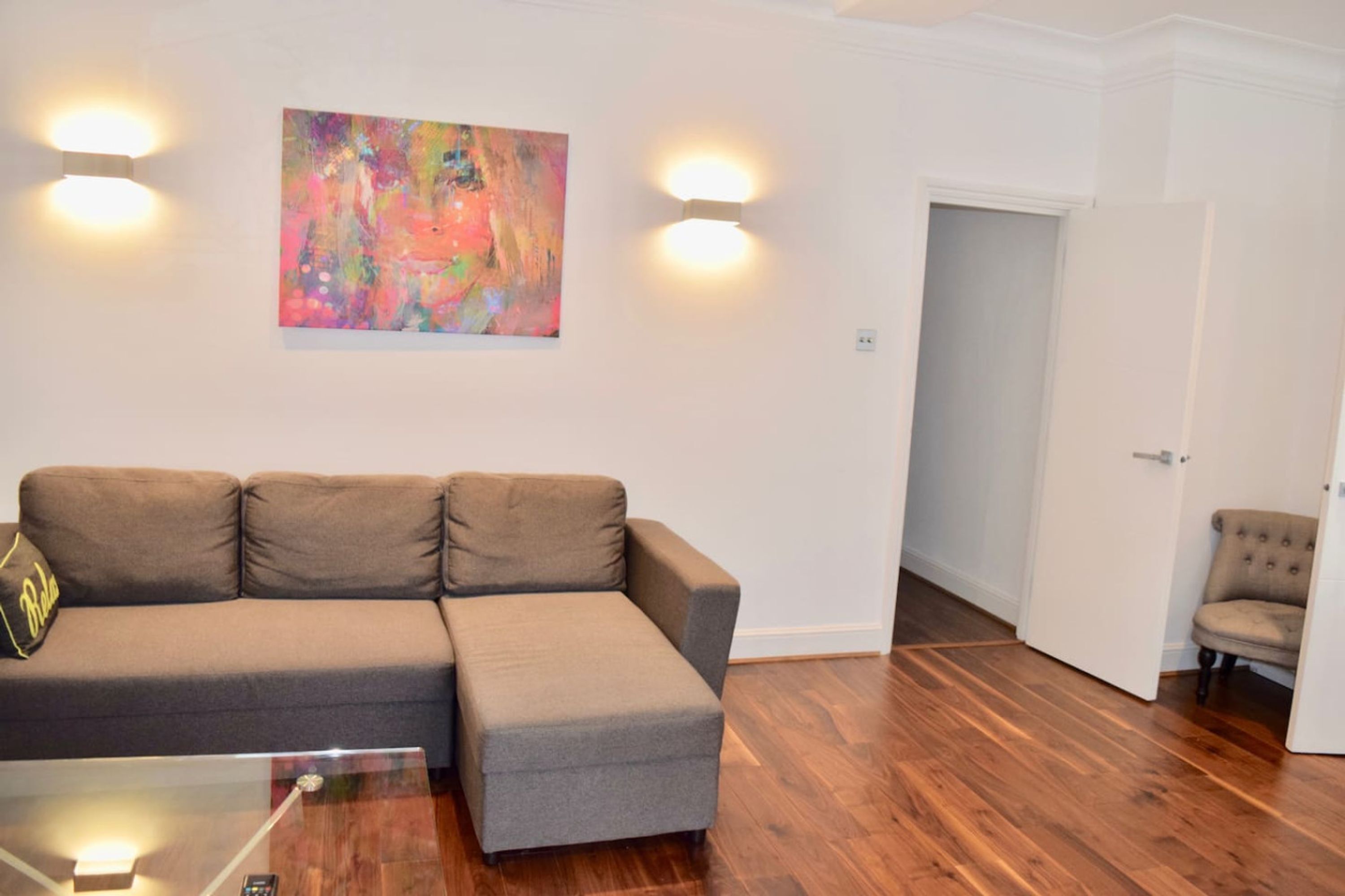 Een woonkamer met een hoekbank, een glazen salontafel en een kleurrijk abstract schilderij aan de muur.