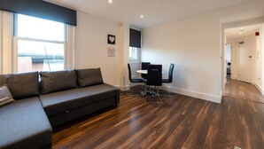 Ein modernes Wohnzimmer mit dunklem Holzboden, einem grauen Sofa und einem Essbereich mit weißem Tisch und schwarzen Stühlen.