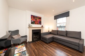 A living room with a sectional sofa, a fireplace, a painting, and a coffee table with magazines. - Stunning Hyde Park Apartment SS3 (London)
