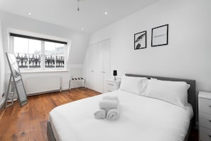 A modern hotel room with a large bed, white linens, a view through a large window, a radiator, and a small bedside table with a lamp. - Central London 3 Bed Flat FS4 (London)