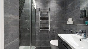 A modern bathroom with a glass shower enclosure, a white toilet, and a large mirror above the sink. - Central London 3 Bed Flat FS4 (London)