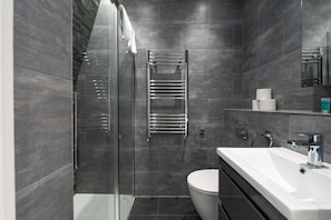 A modern bathroom with a glass shower enclosure, a white toilet, and a large mirror above the sink. - Central London 3 Bed Flat FS4 (London)