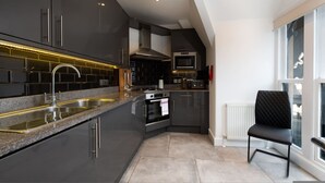 A modern kitchen with dark cabinetry, stainless steel appliances, and a black chair by the window. - Central London 3 Bed Flat FS4 (London)