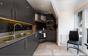 A modern kitchen with dark cabinetry, stainless steel appliances, and a black chair by the window. - Central London 3 Bed Flat FS4 (London)