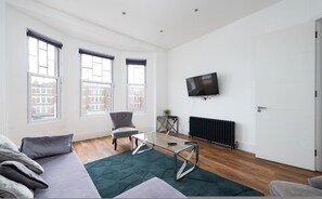 Un salon moderne avec un canapé, une table basse en verre et un fauteuil. De grandes fenêtres offrent une vue sur les bâtiments environnants.