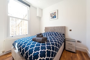 Ein Schlafzimmer mit großem Fenster, einem Bett mit gemustertem Überwurf, einem Nachttisch und einem an der Wand hängenden Bild.