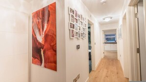 Een gang met een groot abstract schilderij, een reeks ingelijste foto’s aan de muur, en een keuken die zichtbaar is door een open deur.