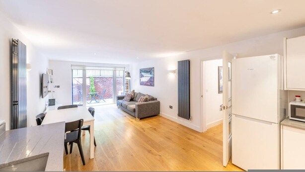 Een moderne keuken met witte apparaten, een eethoek met tafel en stoelen, en een grijze bank in een lichte woonruimte met houten vloeren en grote ramen.